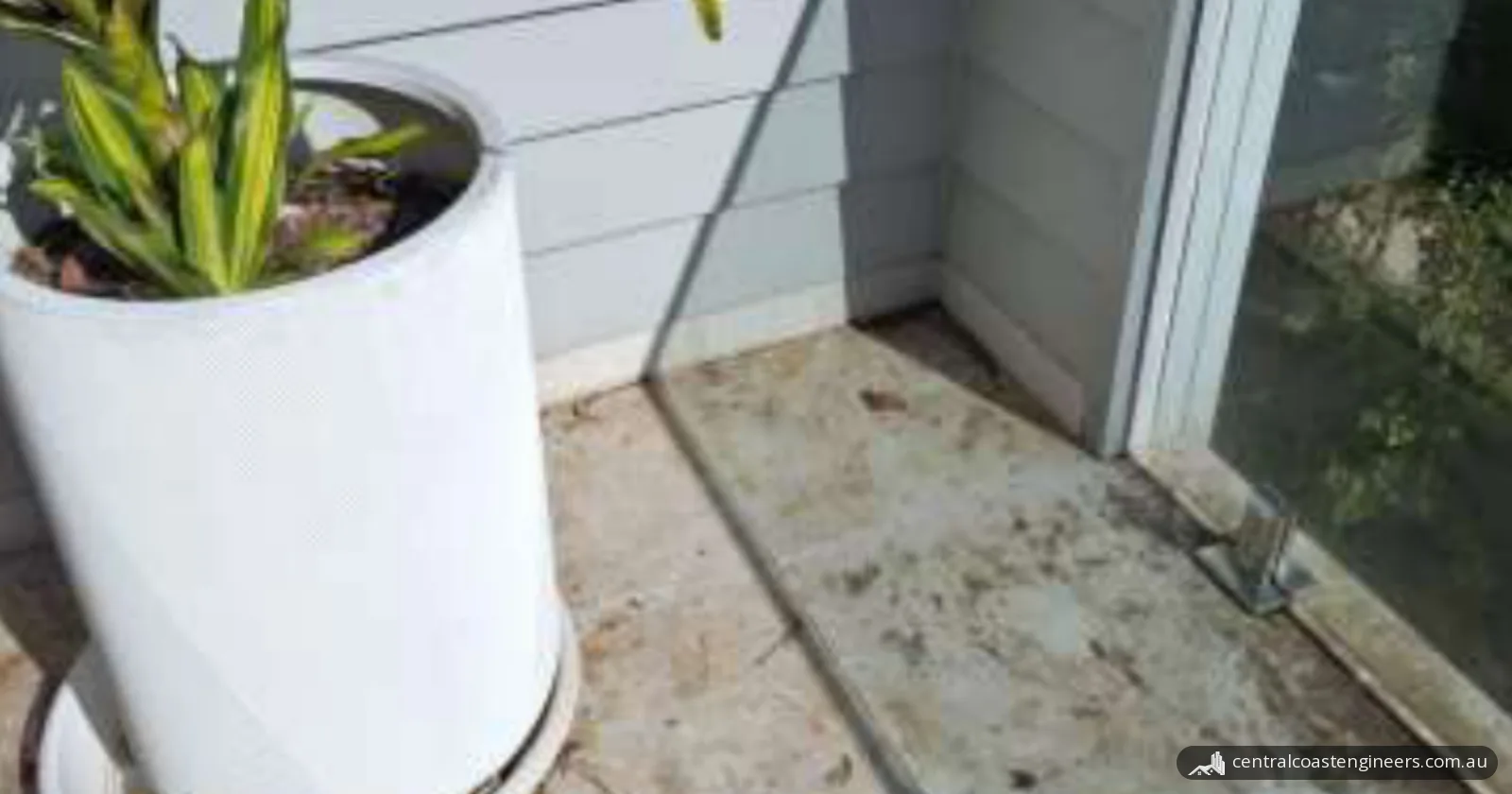 Outer corner of an elevated tiled deck, with a white pot plant against painted cladding, a glass balustrade on metal spigots and stained tiles